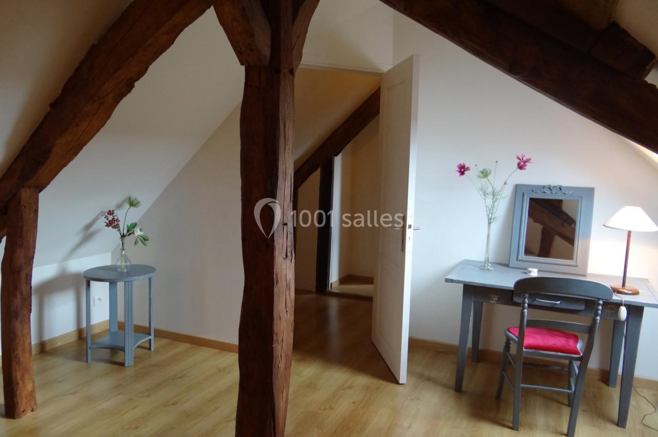 Pièce mansardée avec poutres apparentes, un bureau, une chaise, une lampe et des fleurs sur des meubles en bois.