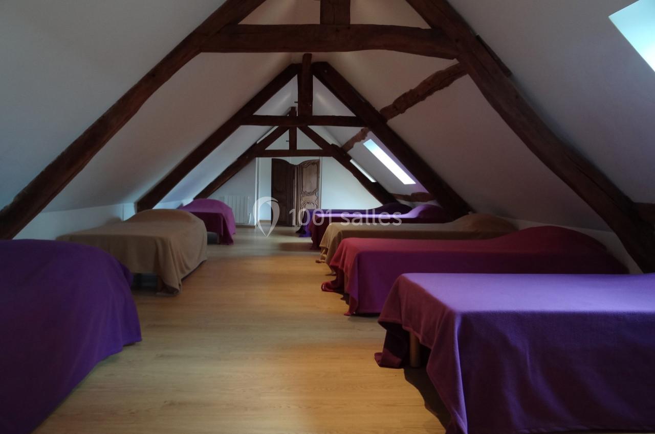 Dortoir mansardé avec poutres apparentes, lits alignés recouverts de couvertures colorées, sol en bois clair.