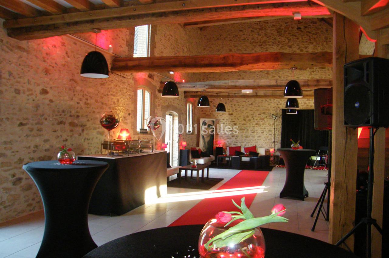 Salle en pierre avec poutres apparentes, tables hautes décorées de fleurs, éclairage rouge et mobilier moderne.