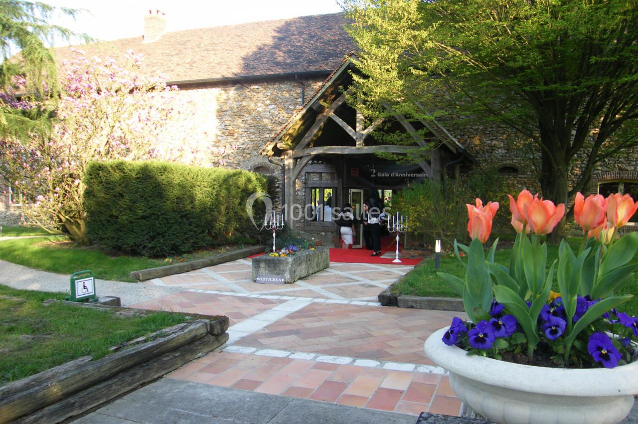 Entrée d'un bâtiment en pierre avec un tapis rouge, entourée de verdure, de fleurs et d'arbres en arrière-plan.