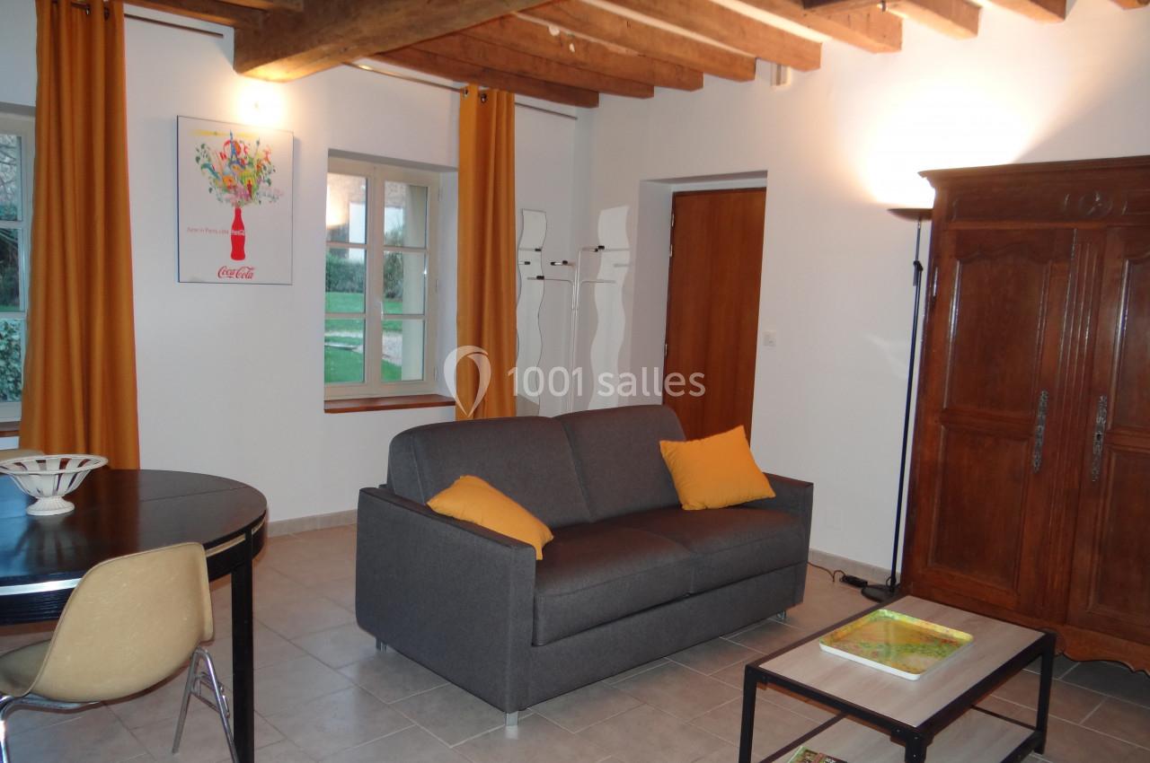 Salon lumineux avec canapé gris, table basse, table à manger, poutres apparentes et rideaux jaunes.