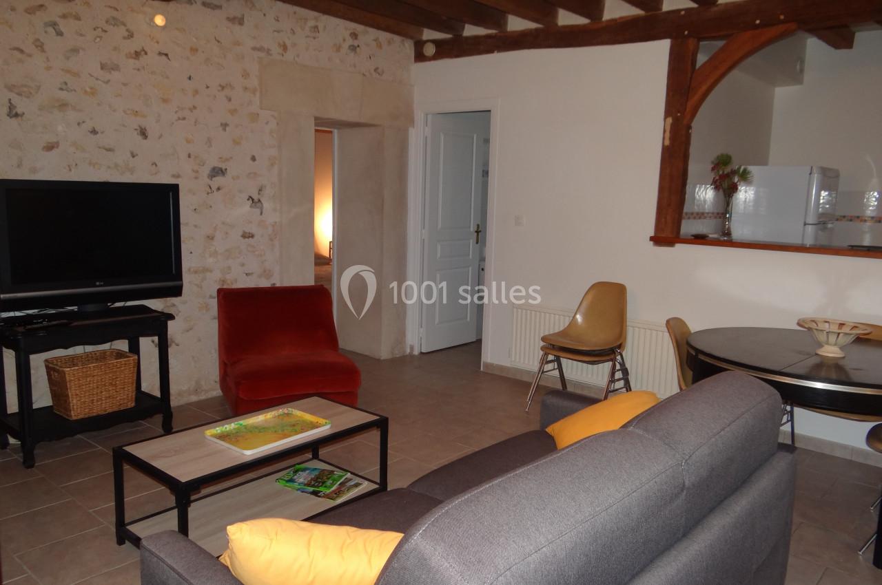 Salon avec murs en pierre, canapé gris, fauteuil rouge, table ronde, télévision et cuisine ouverte en arrière-plan.