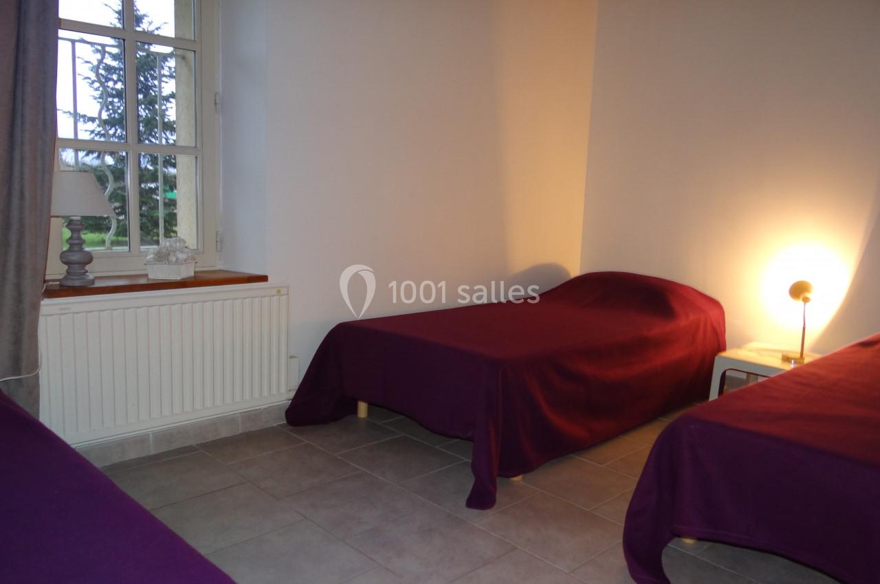 Deux lits simples avec couvre-lits violets dans une chambre lumineuse avec fenêtre, lampe de chevet et sol carrelé.