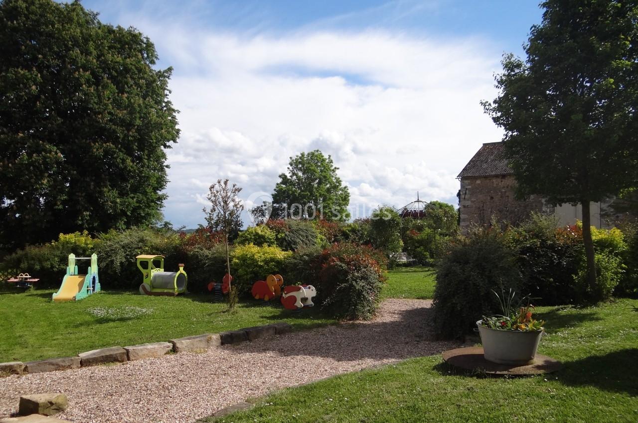 Jardin avec pelouse, jeux pour enfants, arbres et arbustes, sous un ciel bleu partiellement nuageux.