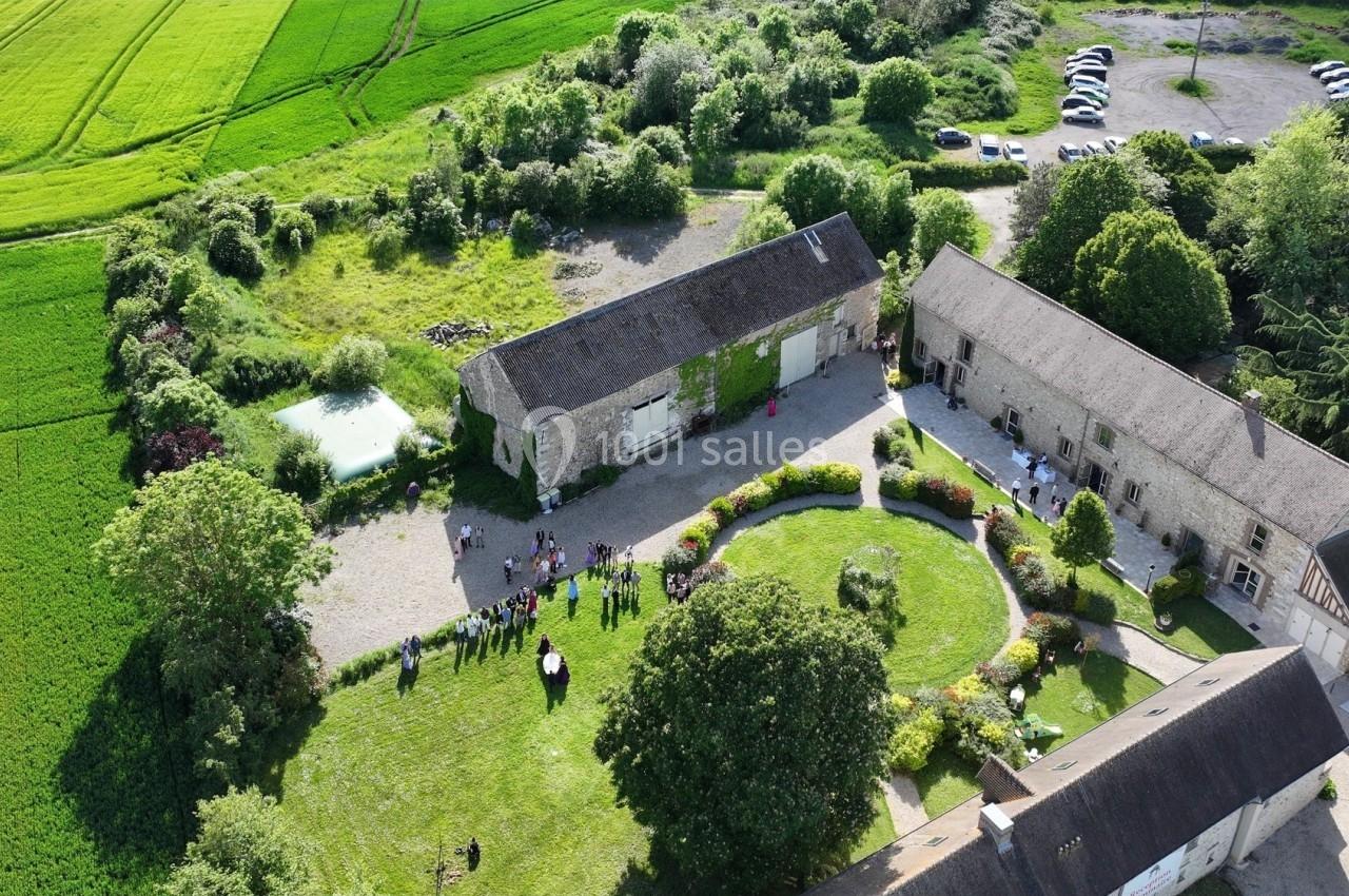 Vue aérienne d'une ferme avec bâtiments en pierre, cour centrale, pelouse, jardins et champs environnants.