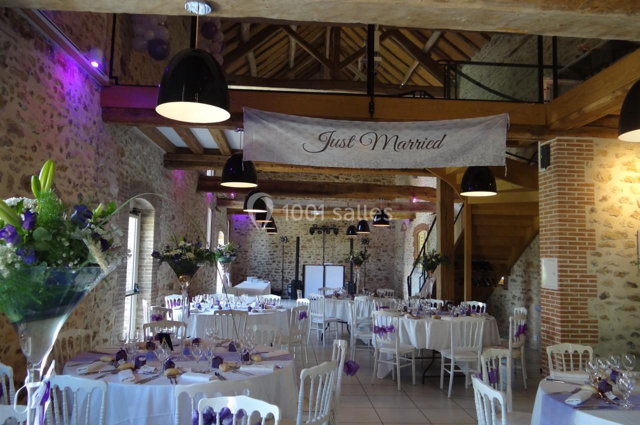 Salle de réception décorée pour un mariage, avec tables dressées, fleurs violettes et une bannière ’Just Married’.