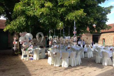 Cour extérieure éclairée en soirée, avec un grand arbre central, des tables, des palettes en bois et des invités debout.