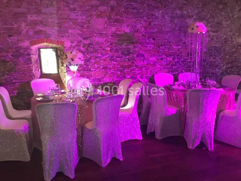 Salle décorée pour un événement avec des tables rondes, nappes scintillantes, chaises blanches et éclairage rose.