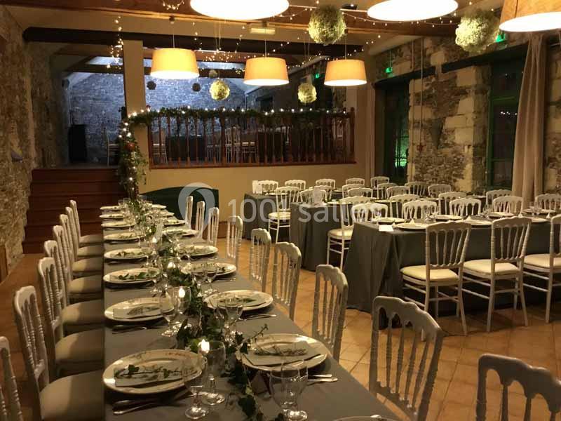 Salle de réception décorée avec des tables alignées, nappes grises, chaises blanches et guirlandes lumineuses.