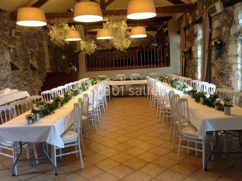 Salle de réception avec tables rectangulaires décorées de nappes blanches et de feuillages, éclairée par des suspensions.