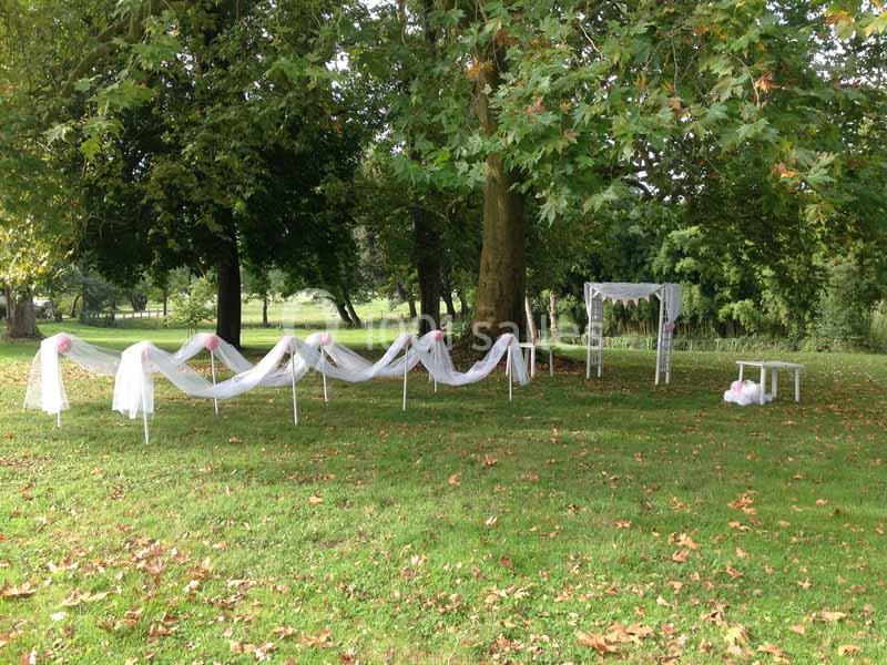 Décoration de mariage en extérieur avec arche florale, voilages blancs et chaises sur une pelouse entourée d'arbres.