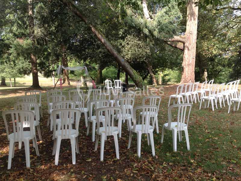 Chaises blanches en plastique disposées en rangées sur une pelouse ombragée, entourées d'arbres.