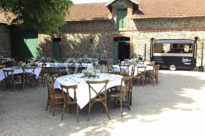 Cour extérieure éclairée en soirée, avec un grand arbre central, des tables, des palettes en bois et des invités debout.