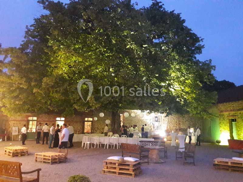 Cour extérieure éclairée en soirée, avec un grand arbre central, des tables, des palettes en bois et des invités debout.