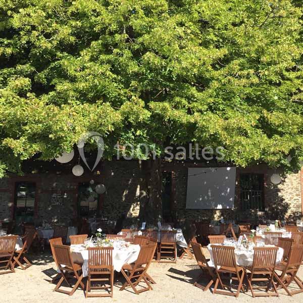 Tables dressées avec nappes blanches et chaises en bois disposées sous un grand arbre dans une cour ensoleillée.