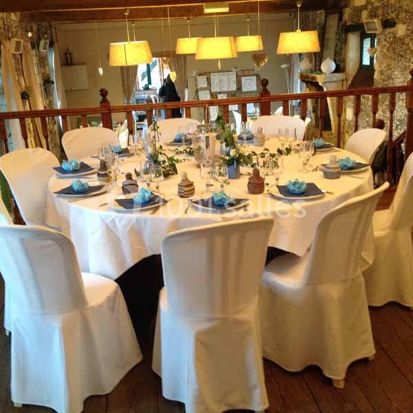 Table ronde élégamment dressée avec nappes blanches, serviettes bleues et décorations, dans une salle lumineuse en bois.