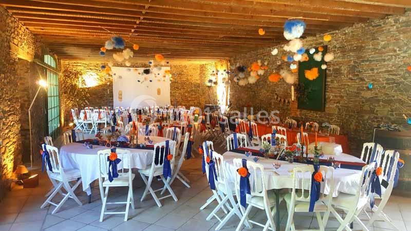 Salle en pierre décorée pour un événement, avec tables rondes dressées, chaises blanches et suspensions colorées.