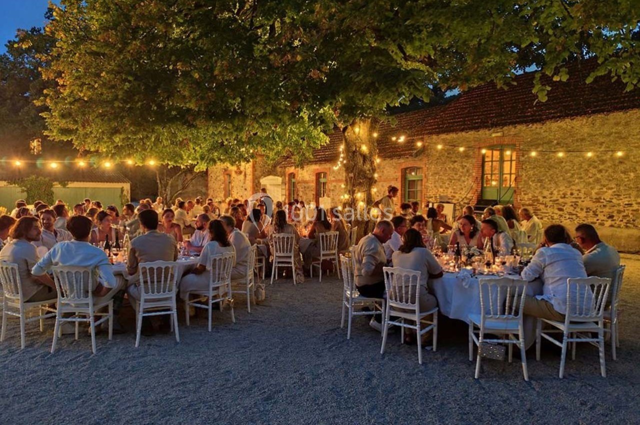 Dîner en plein air sous des guirlandes lumineuses, avec des invités assis autour de tables rondes près d'un bâtiment en…