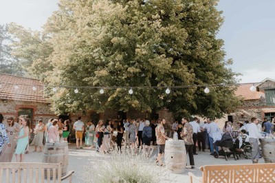 Un groupe de personnes rassemblées sous de grands arbres devant des bâtiments en pierre par une journée ensoleillée.