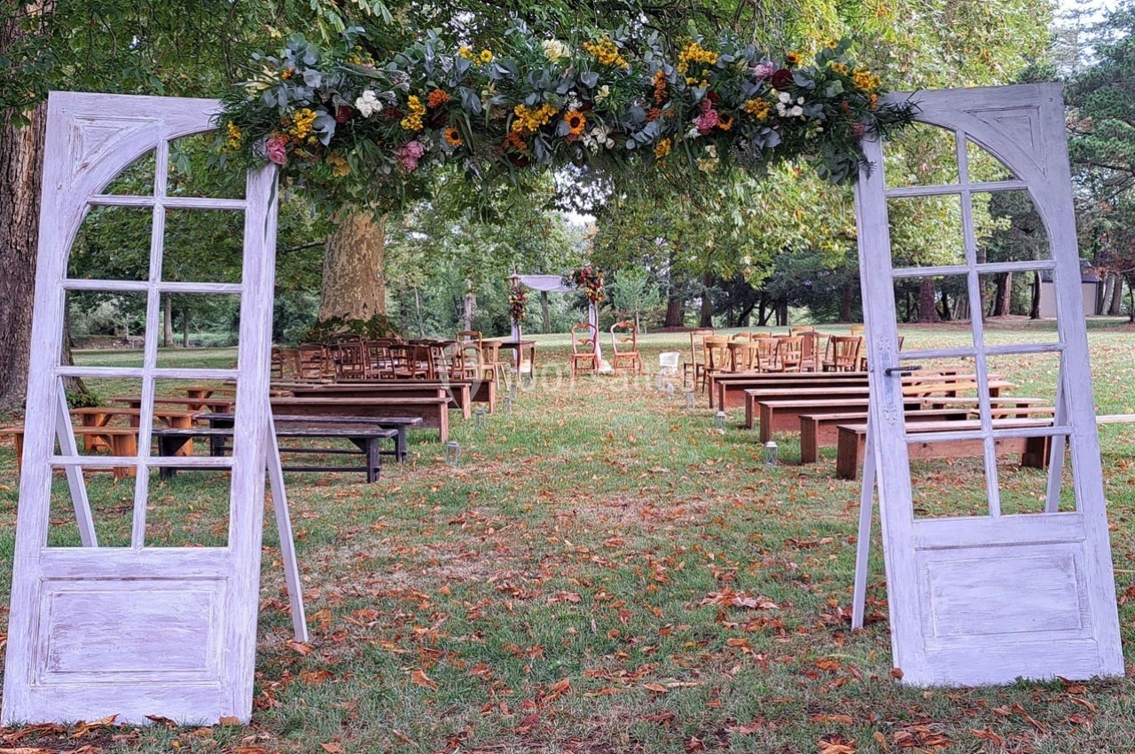 Allée de cérémonie en plein air avec bancs en bois, encadrée par une arche décorée de fleurs et de portes blanches.