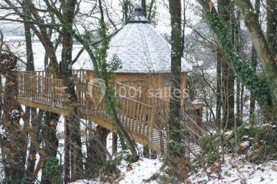 Cabane en bois perchée dans les arbres avec une passerelle en bois, entourée de végétation automnale et deux vélos au…