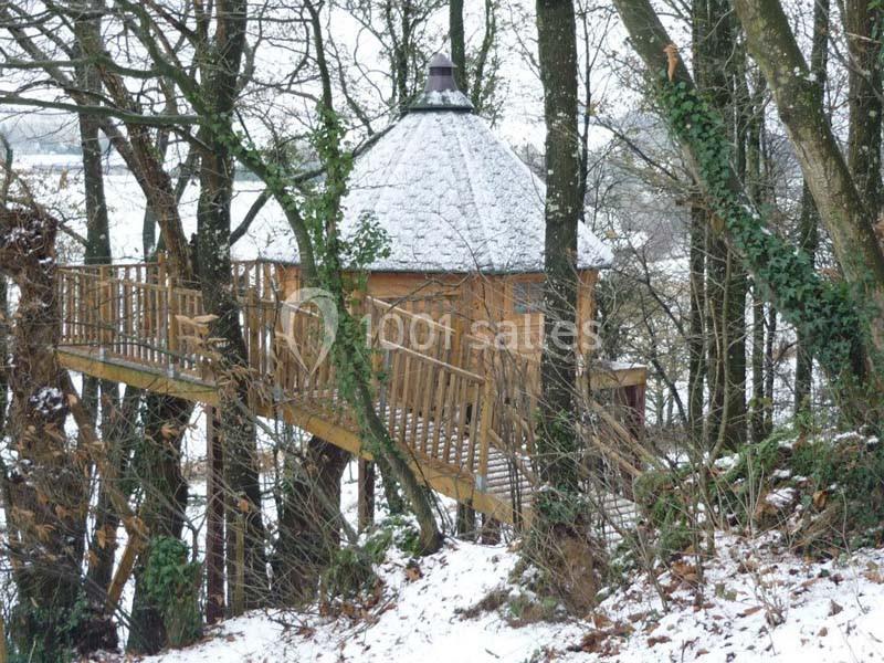 Cabane en bois perchée dans les arbres, entourée de neige et accessible par une passerelle en bois.
