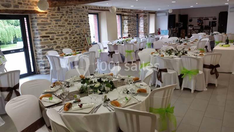Salle de réception décorée avec des tables rondes dressées, nappes blanches et chaises ornées de rubans verts.
