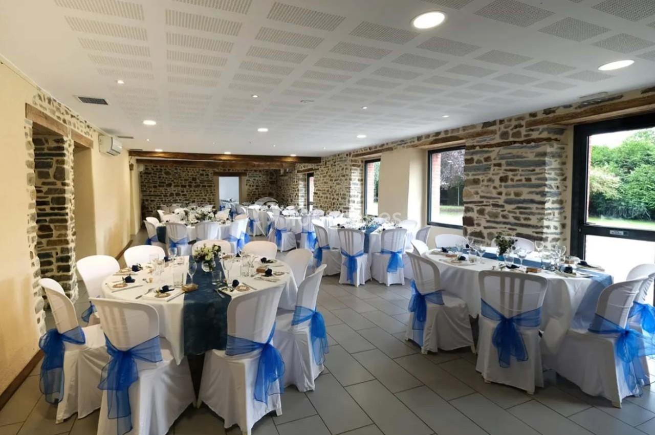 Salle de réception décorée avec des tables rondes, nappes blanches et nœuds bleus sur les chaises.