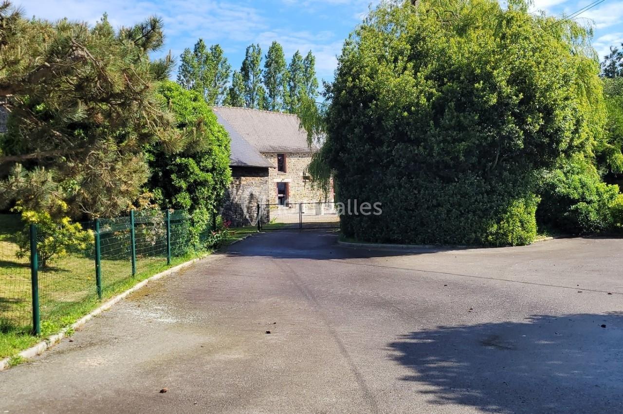 Entrée d'un domaine avec allée asphaltée, bâtiments en pierre, arbres et panneaux indiquant le parking et la réception.