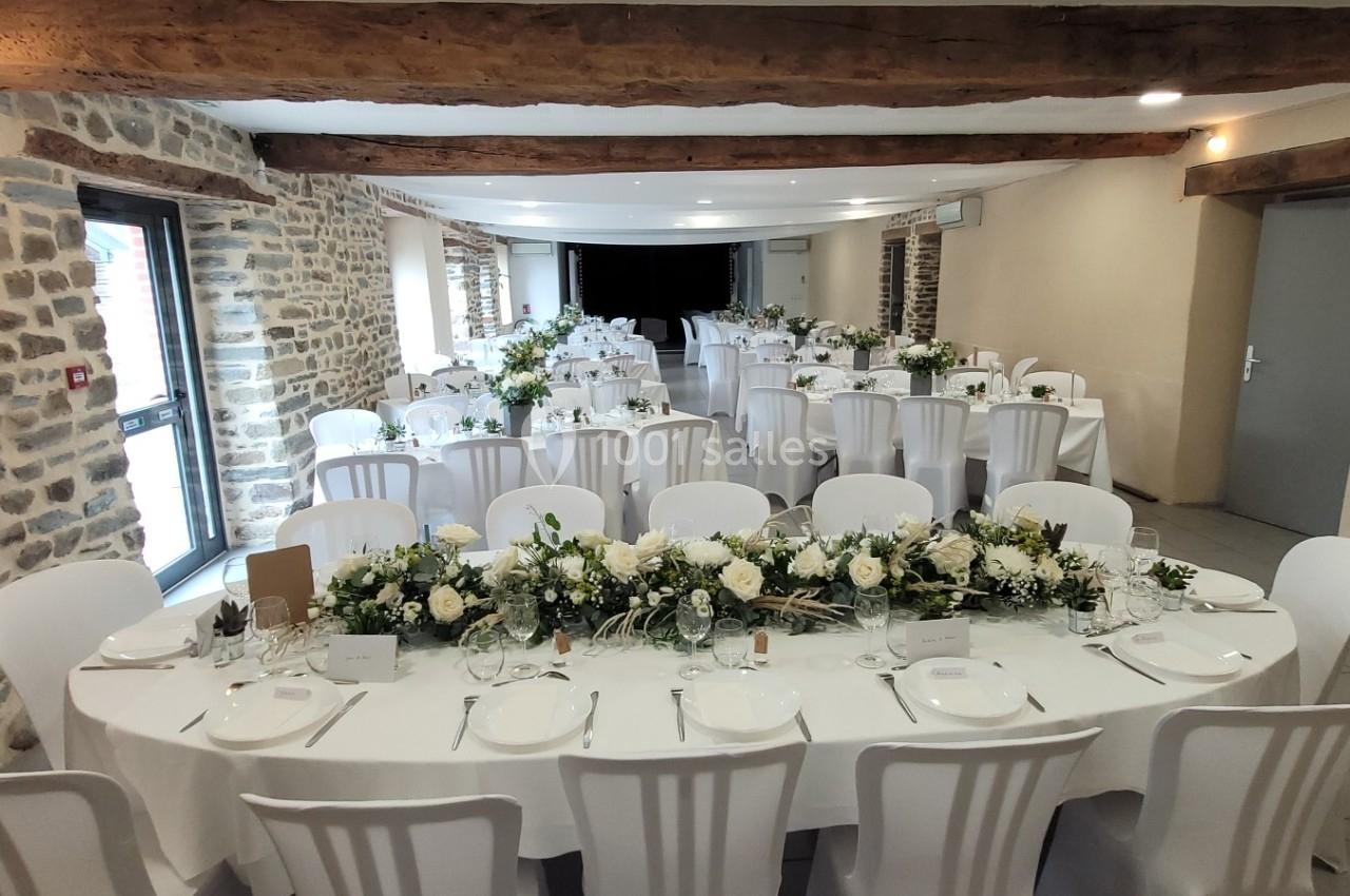 Salle de réception avec tables dressées, nappes blanches, chaises blanches et décorations florales.