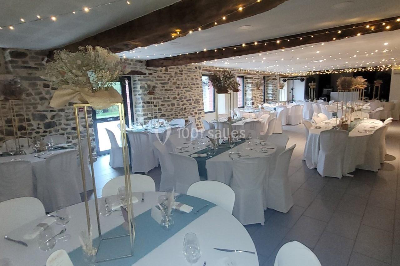 Salle de réception décorée avec des tables rondes et rectangulaires, nappes blanches et guirlandes lumineuses suspendues.