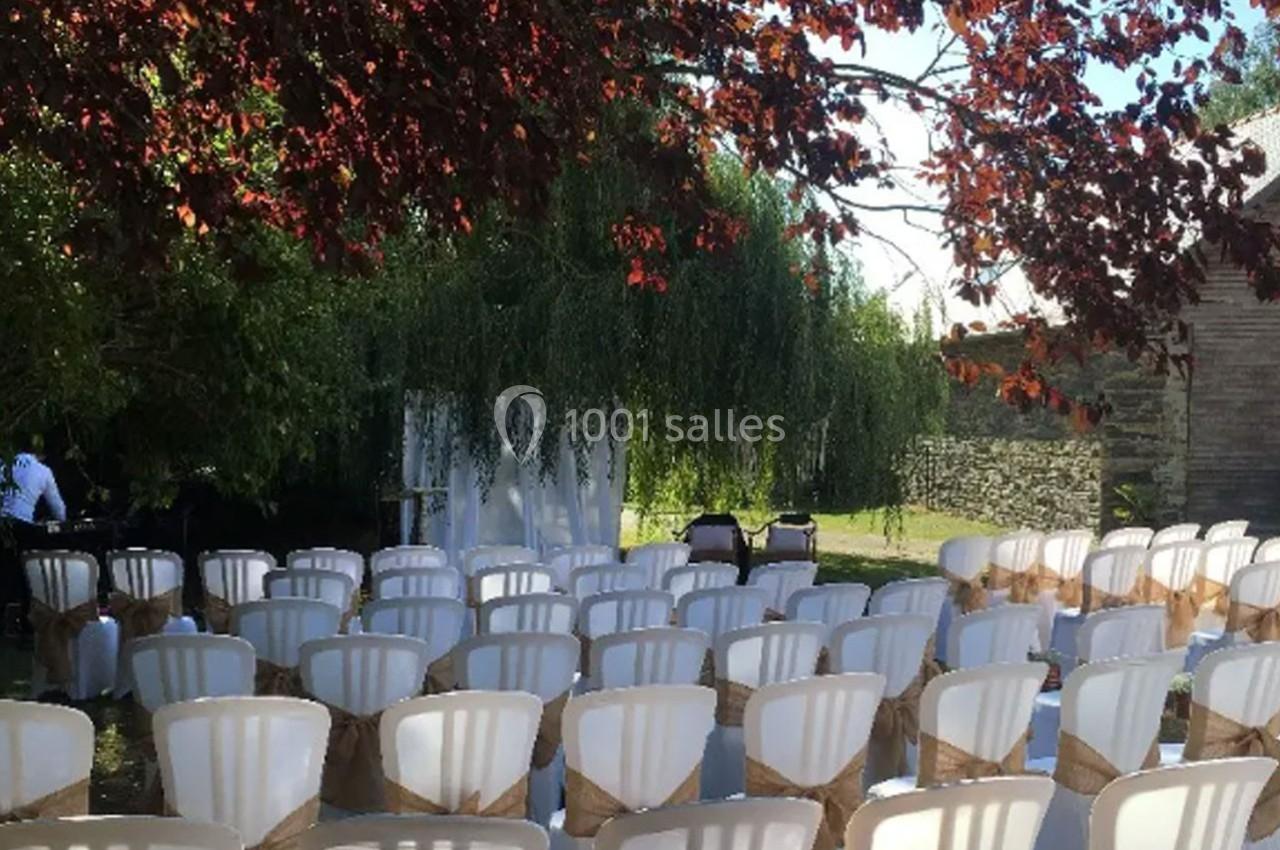 Chaises blanches alignées en extérieur sous des arbres, décorées de rubans beige, prêtes pour une cérémonie.
