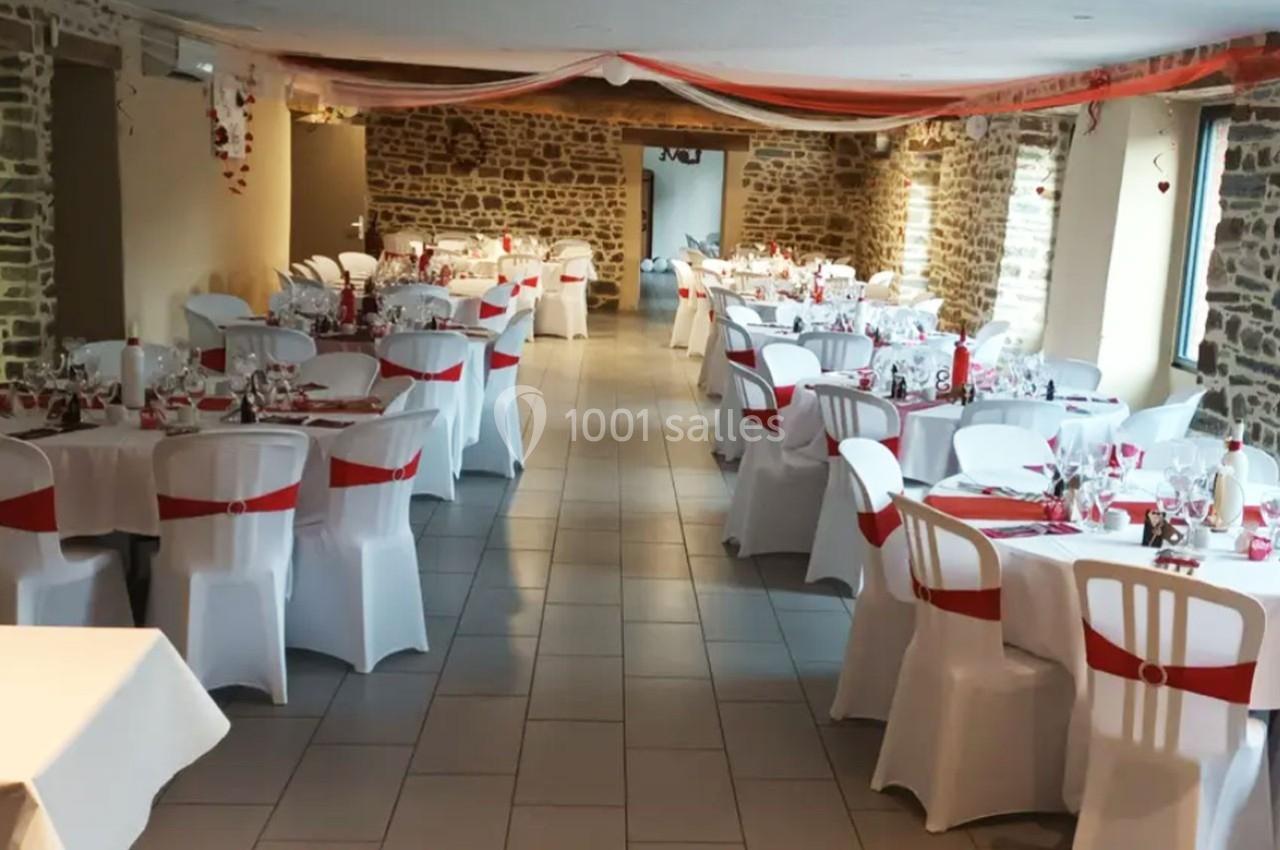 Salle de réception décorée avec des tables rondes, nappes blanches et accents rouges, murs en pierre apparente.