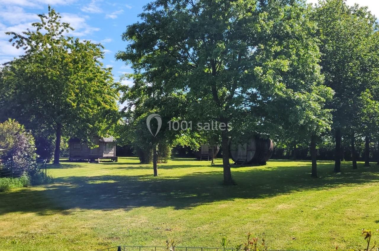 Un jardin verdoyant avec pelouse, arbres et cabanes en bois sous un ciel bleu partiellement nuageux.