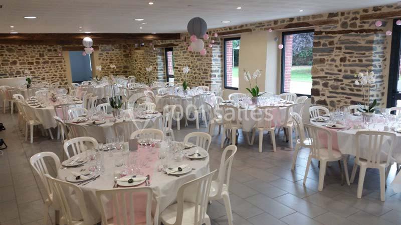 Salle de réception décorée avec des tables rondes dressées, nappes blanches et touches de rose, murs en pierre apparente.