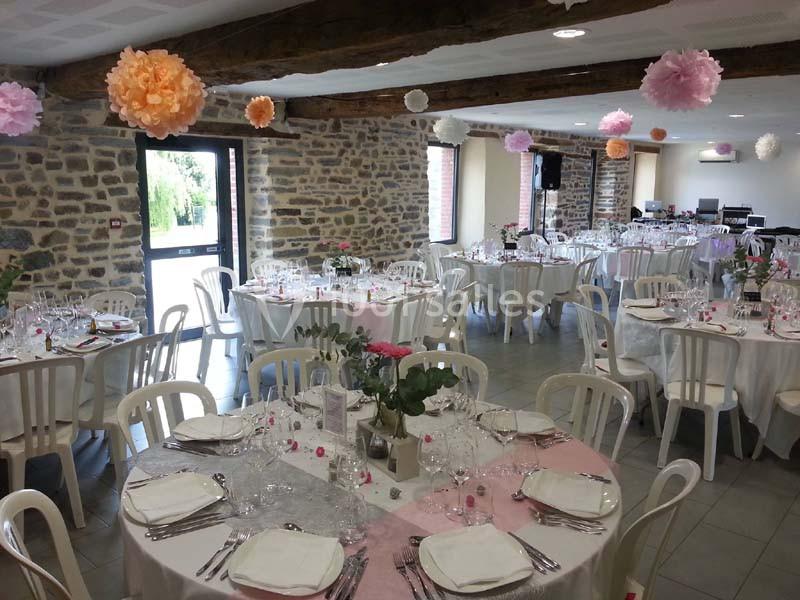 Salle de réception décorée avec des tables rondes dressées, nappes roses et blanches, et suspensions florales colorées.