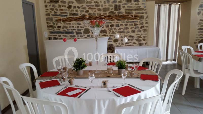 Table ronde dressée avec nappes blanches, serviettes rouges, verres et décorations végétales dans une salle en pierre…