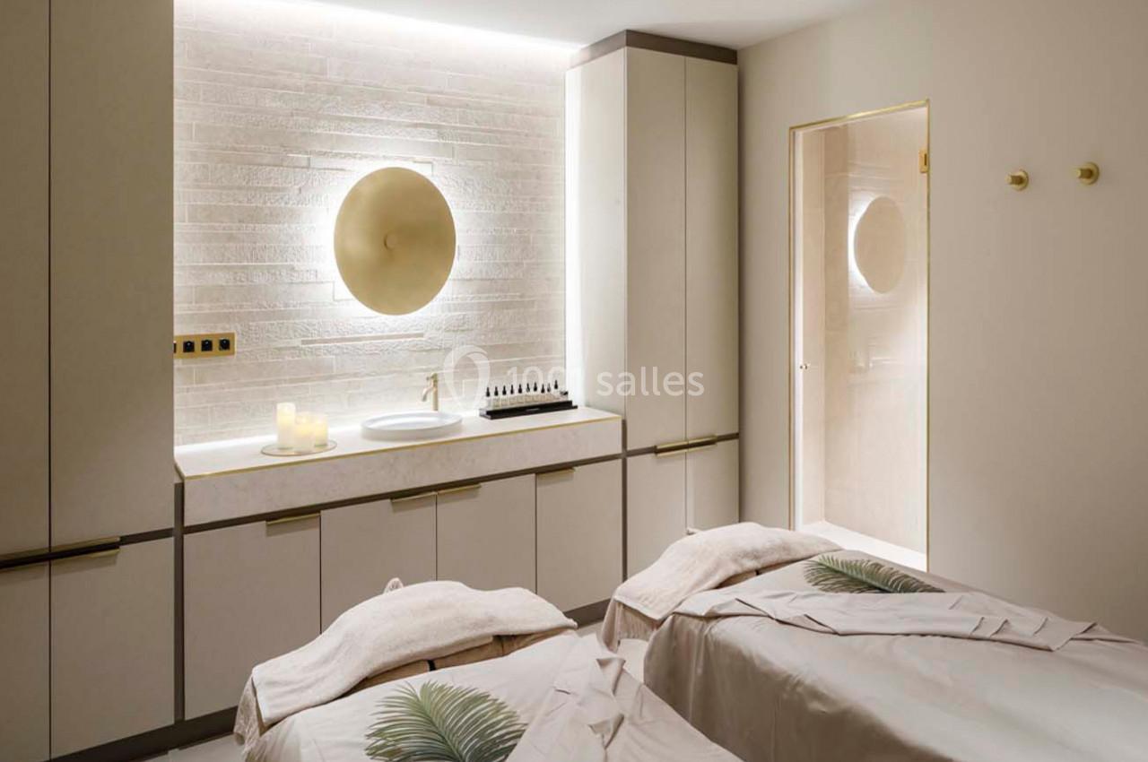 Salle de soins avec deux tables de massage couvertes de draps blancs, éclairage doux et décor minimaliste.