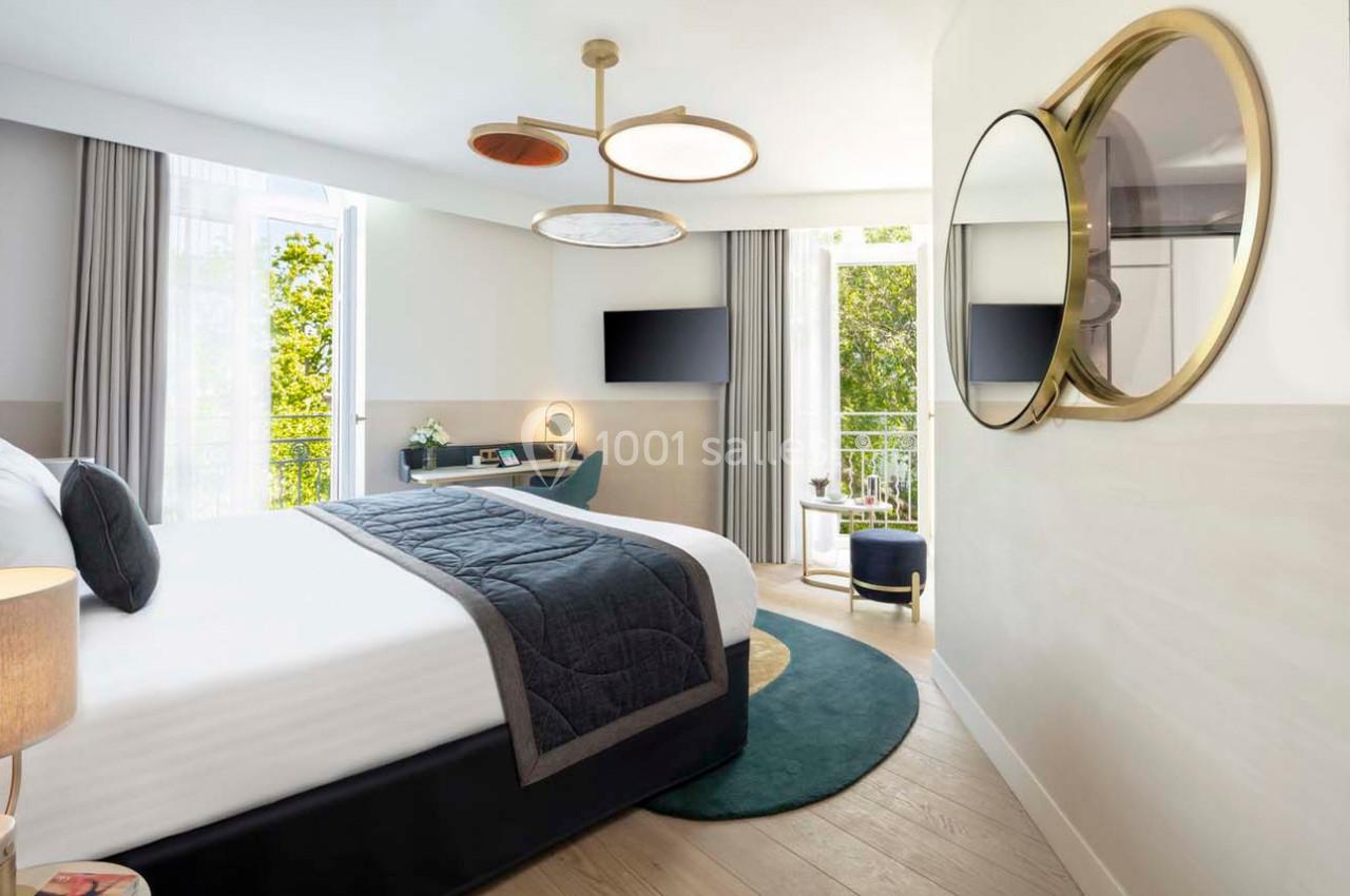 Chambre lumineuse avec lit double, bureau, télévision, miroir rond et balcon donnant sur des arbres.