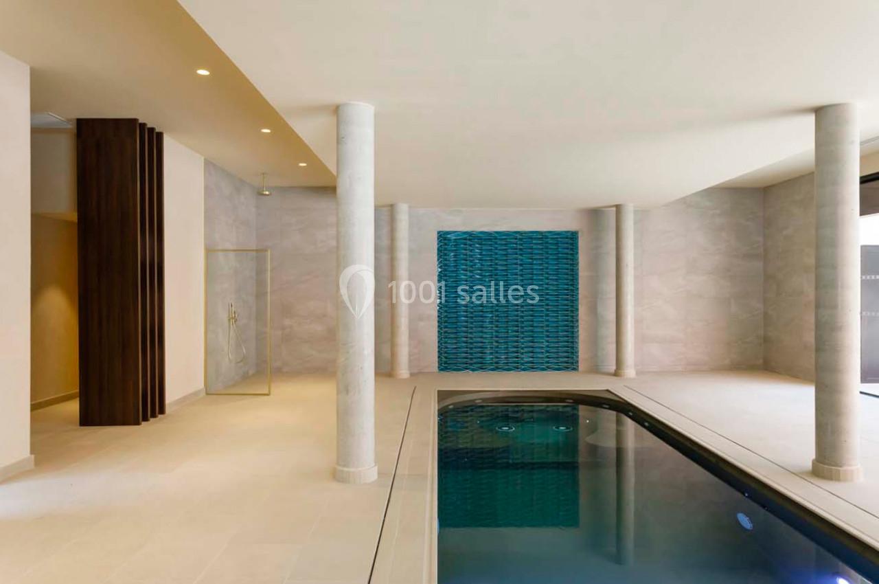 Piscine intérieure moderne avec carrelage beige, mur décoratif bleu et colonnes blanches.