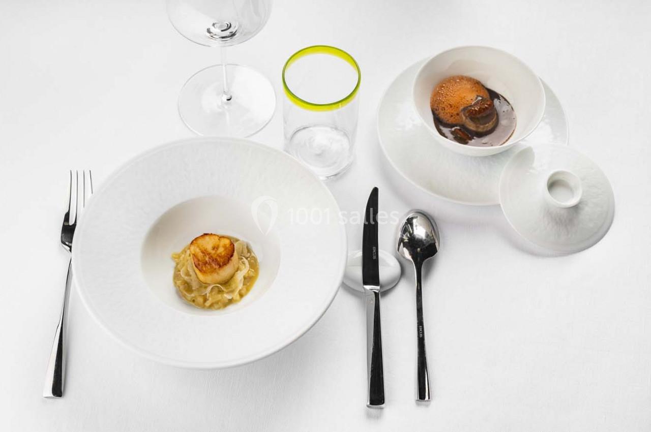 Assiette gastronomique avec ravioles et noix de Saint-Jacques, accompagnée d'une soupe et d'ustensiles sur une nappe blanche.