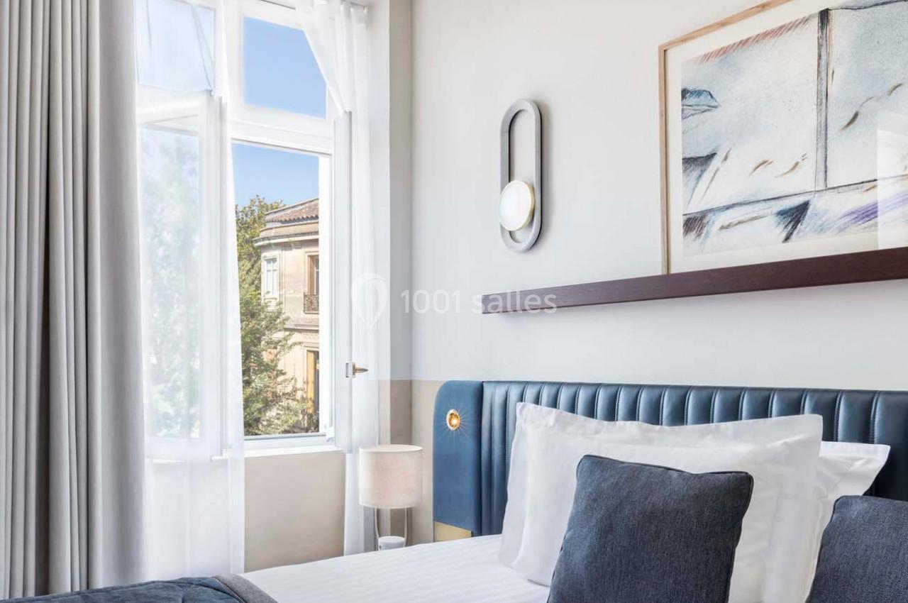 Chambre lumineuse avec lit, coussins bleus, lampe de chevet et grande fenêtre donnant sur un bâtiment ancien.