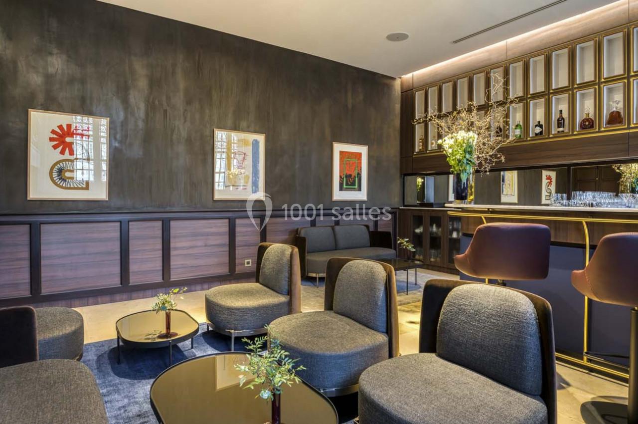 Salon moderne avec fauteuils gris, tables basses dorées, bar en arrière-plan et décorations murales colorées.