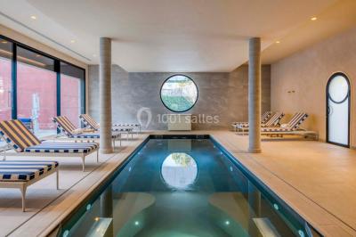 Piscine intérieure avec chaises longues rayées, éclairage doux et grande fenêtre ronde donnant sur l'extérieur.