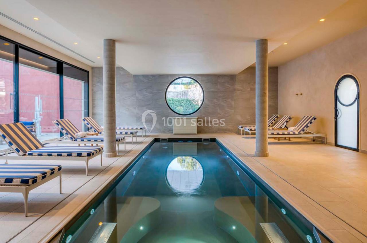 Piscine intérieure avec chaises longues rayées, éclairage doux et grande fenêtre ronde donnant sur l'extérieur.