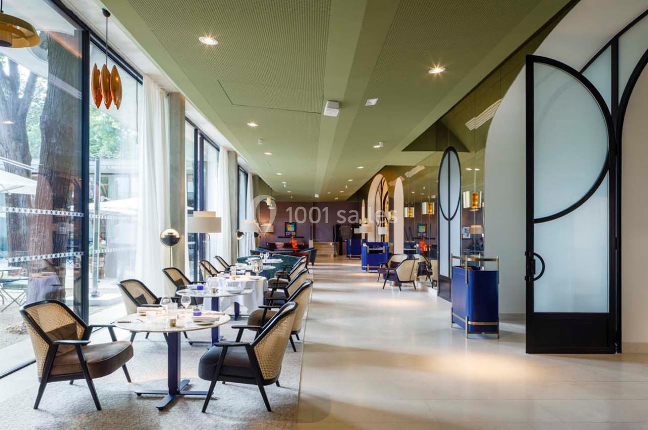 Salle lumineuse avec tables dressées, chaises en rotin, grandes baies vitrées et décoration moderne.