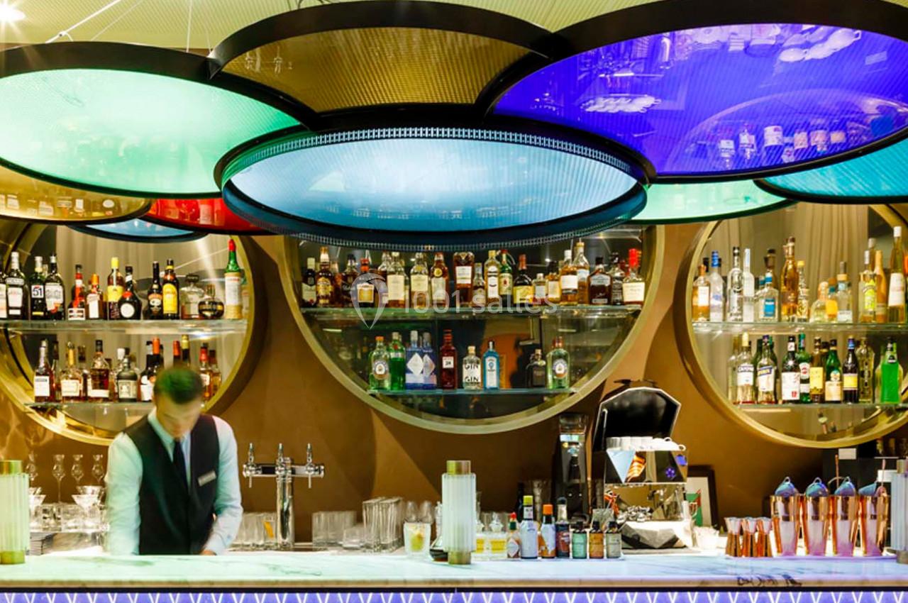 Un barman prépare des boissons derrière un comptoir, avec des étagères remplies de bouteilles et des lumières colorées au…