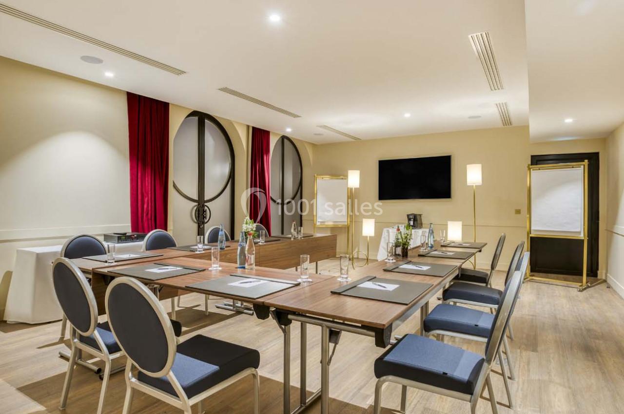 Salle de réunion lumineuse avec tables disposées en U, chaises, écran mural, paperboards et rideaux rouges.