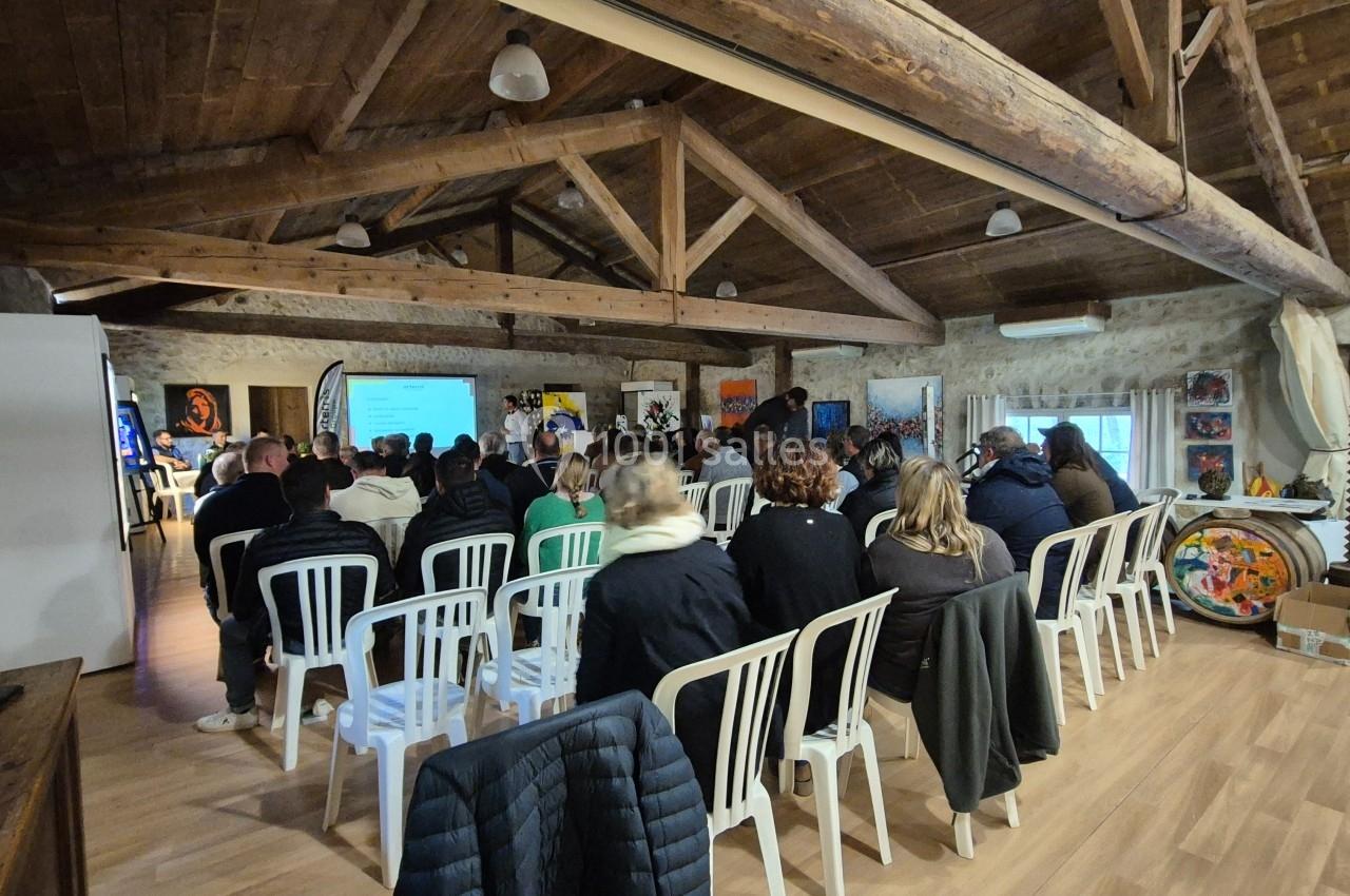 Des personnes assises sur des chaises blanches assistent à une présentation dans une salle avec des poutres en bois…