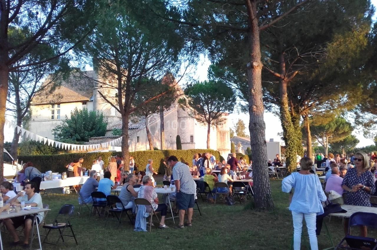 Des personnes assises ou debout participent à un événement en plein air près d'un bâtiment ancien entouré d'arbres.